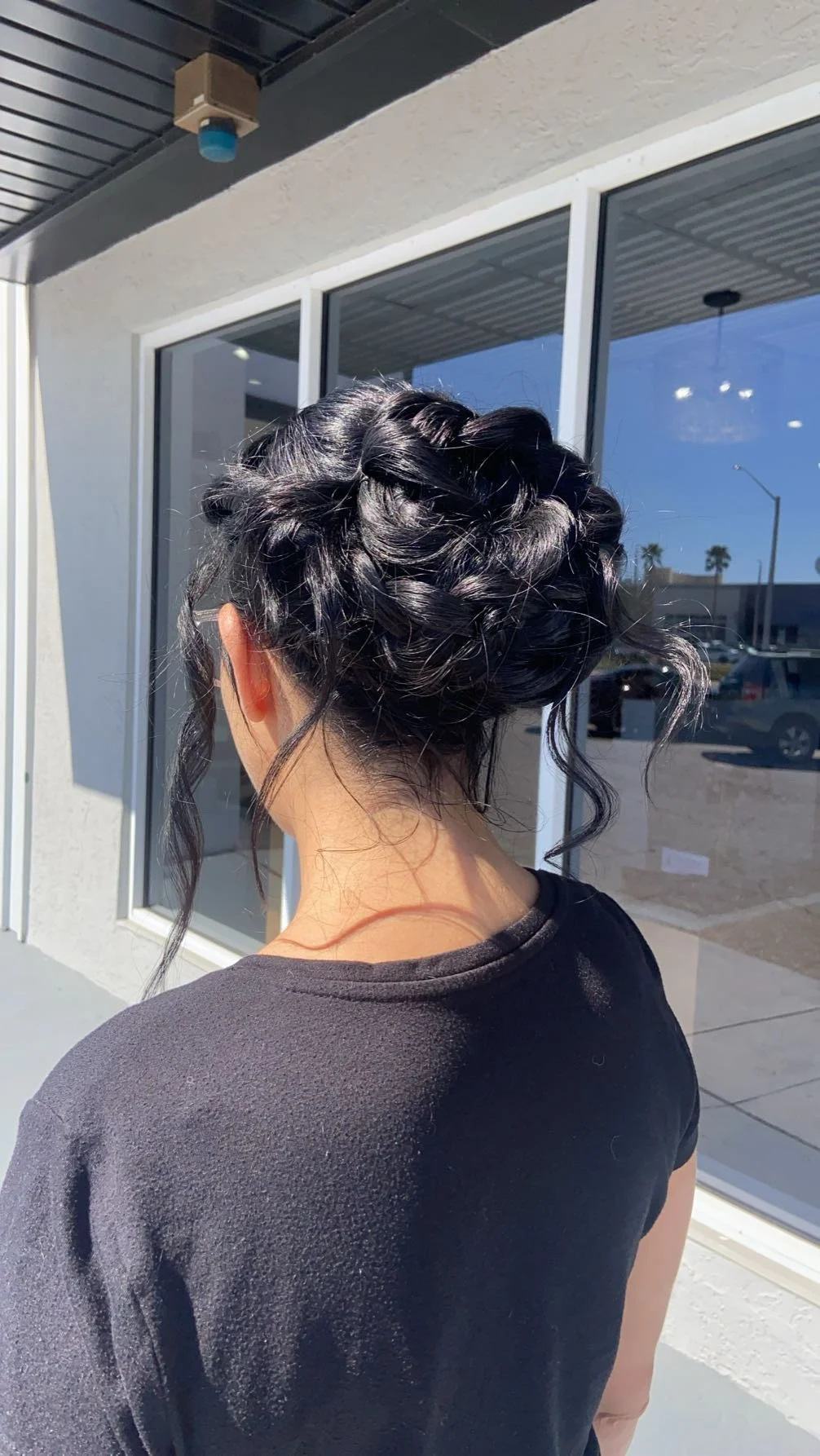 Back view of a woman with dark, curly hair styled in an updo, wearing a black T-shirt, standing outside near a building with large glass windows.