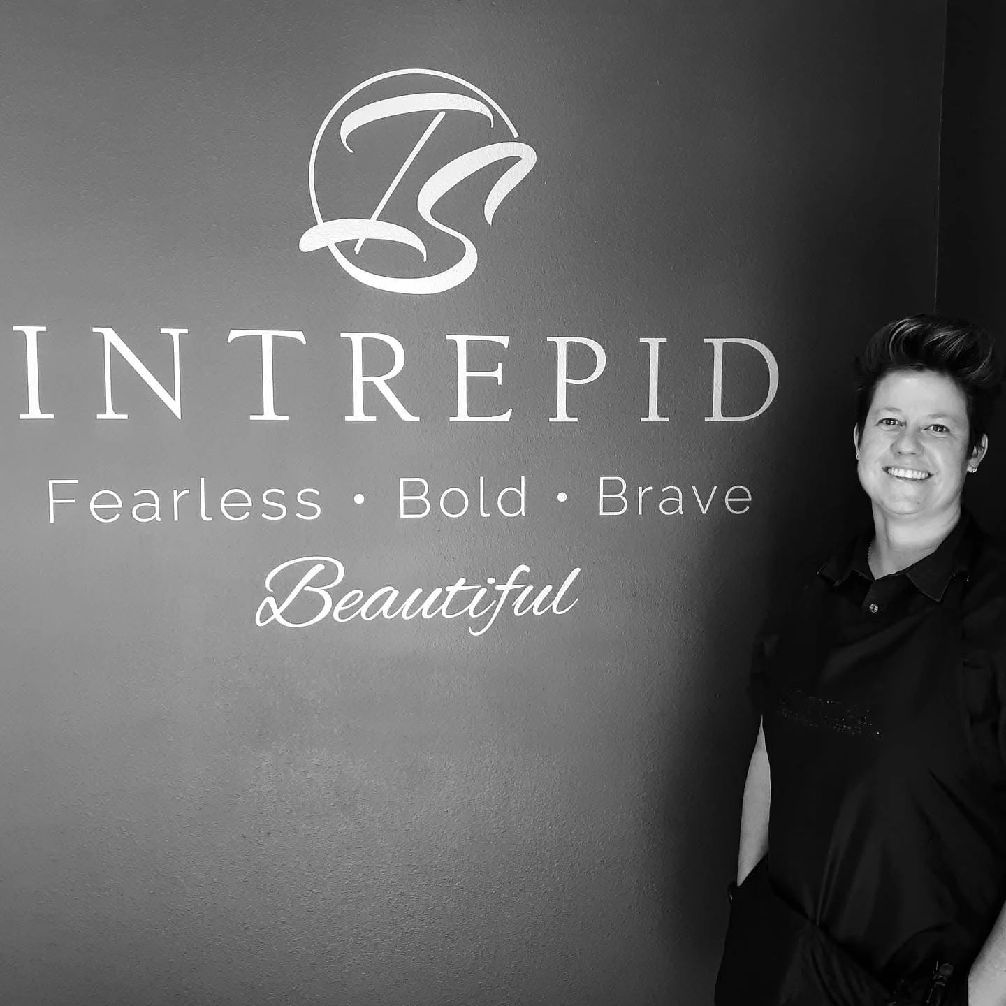A woman smiling standing next to a wall with the logo and words "INTREPID" and the phrase "Fearless • Bold • Brave" in white on a dark background.