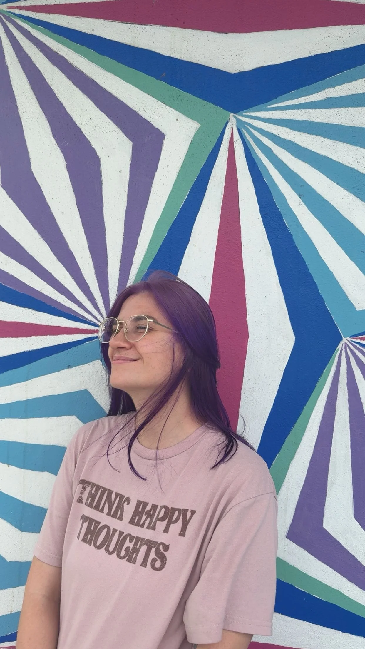 A young woman with dark hair and glasses standing in front of a colorful geometric mural painted on a wall.