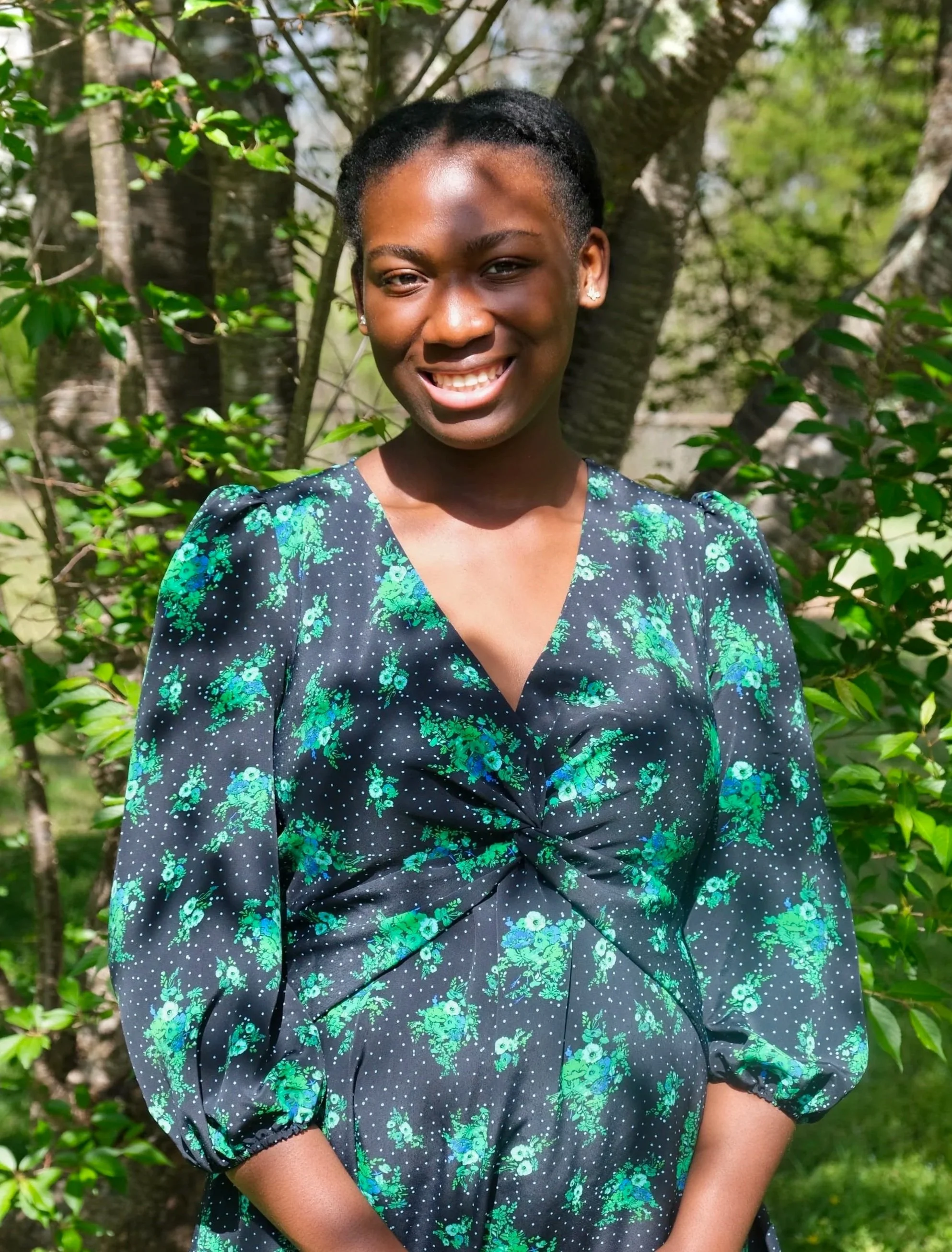 A young woman with dark skin and black hair styled in braids or twists, smiling with her teeth visible, outdoors with green trees and foliage in the background, wearing a dark dress with green floral patterns and puffy sleeves.