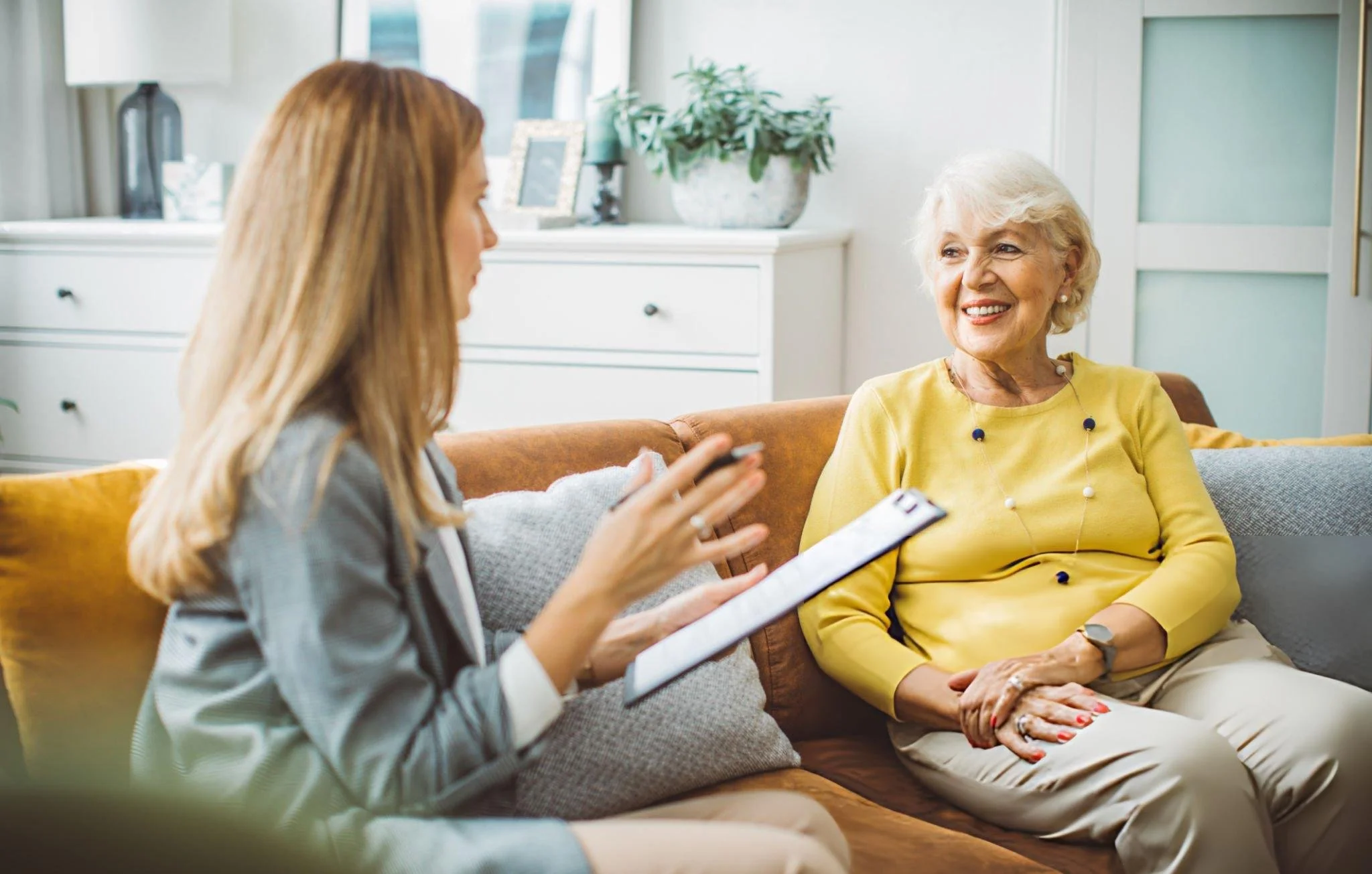 A young woman and an elderly woman sitting on a sofa, engaging in conversation in a bright living room.