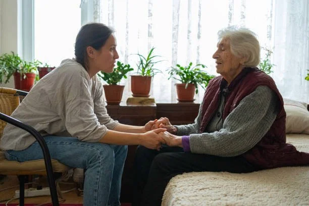 A young woman and an elderly woman sit facing each other, holding hands, in a cozy room with potted plants on the windowsill.