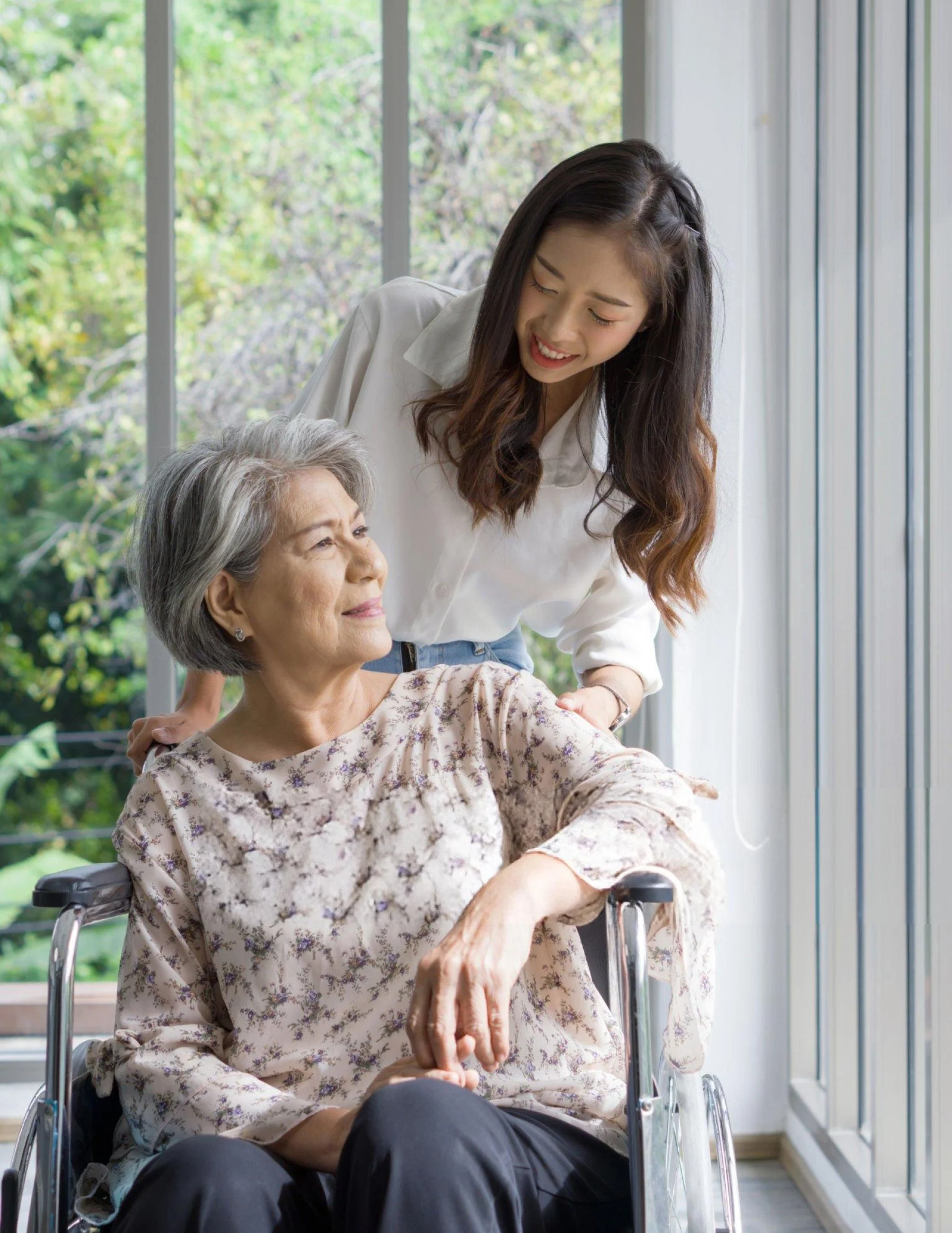A young woman with long dark hair smiling and leaning over an elderly woman with gray hair, sitting in a wheelchair, inside a room with large windows showing greenery outside.