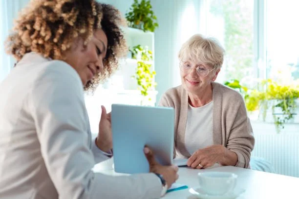 A young woman and an elderly woman sitting at a table, engaging in a conversation, with the young woman holding a clipboard.