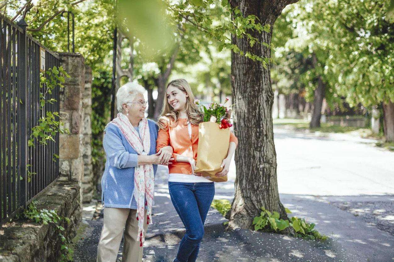A young woman is giving a gift bag with flowers to an elderly woman on a sidewalk under a tree, with a fence and greenery in the background.
