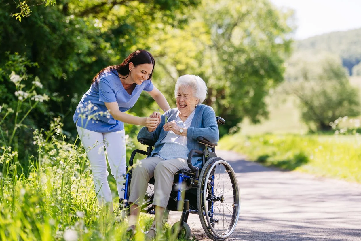 A young woman in medical scrubs helping an elderly woman in a wheelchair outdoors in a green, sunny park.