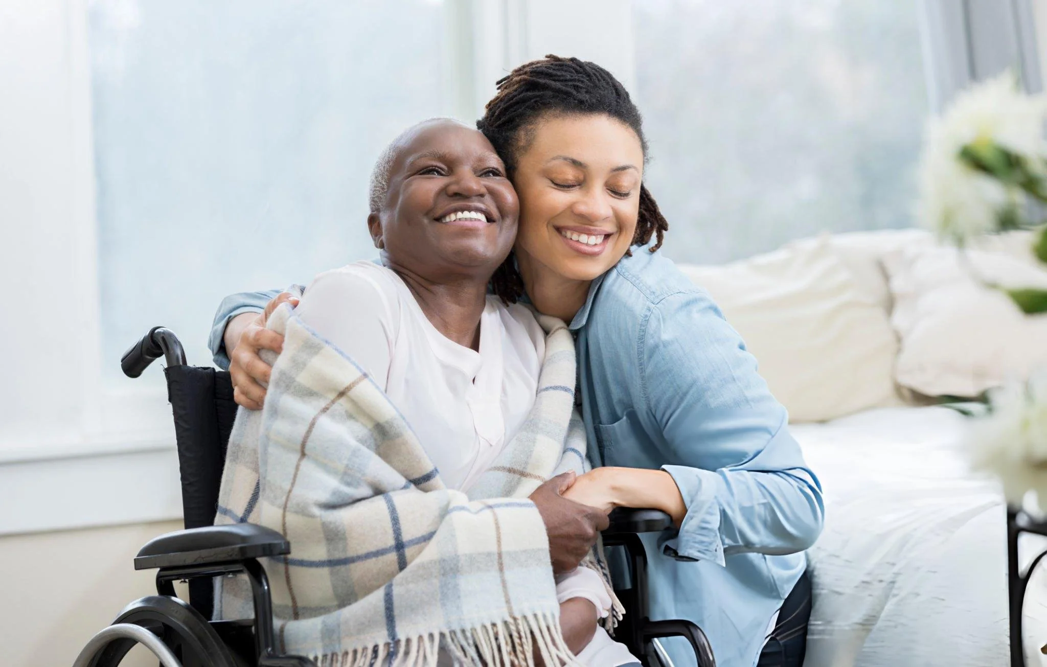A woman in a wheelchair hugging a young woman with dreadlocks, both smiling with eyes closed, in a bright room with large windows.