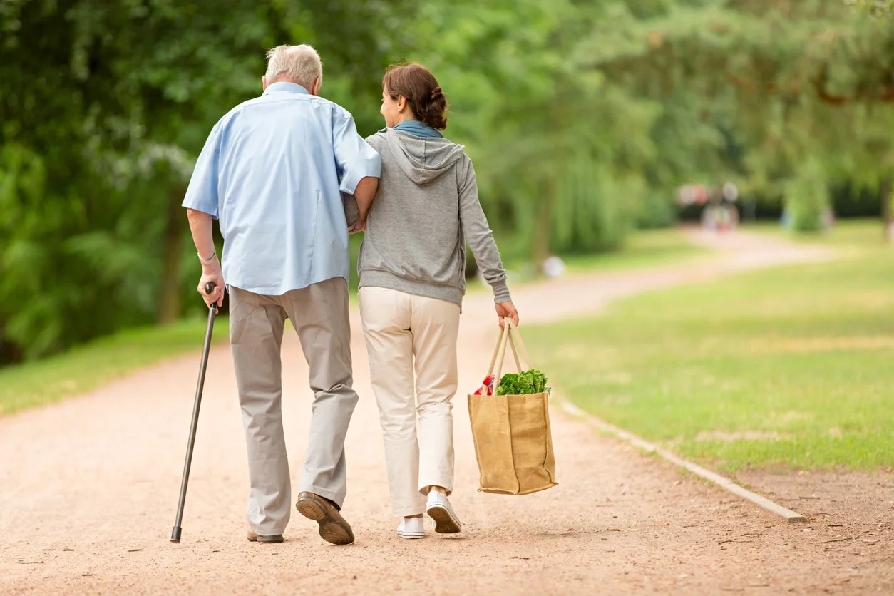 An elderly man and a woman walking together in a park, with the woman carrying a grocery bag.