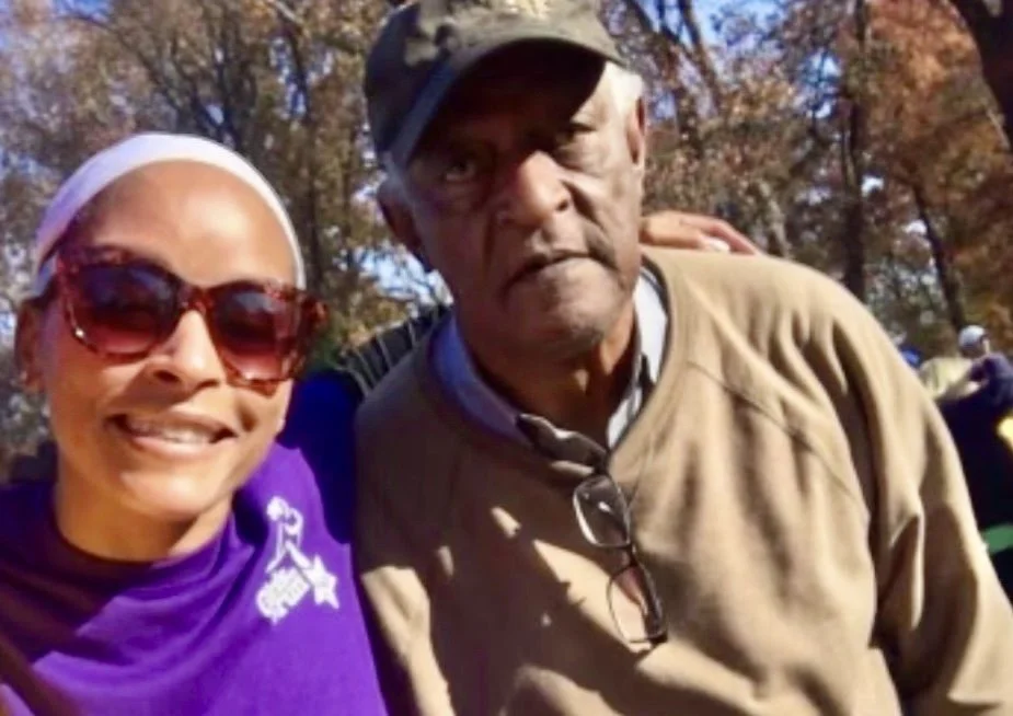 A woman wearing glasses and a purple shirt taking a selfie with an older man wearing a tan sweater and a cap outdoors during fall.