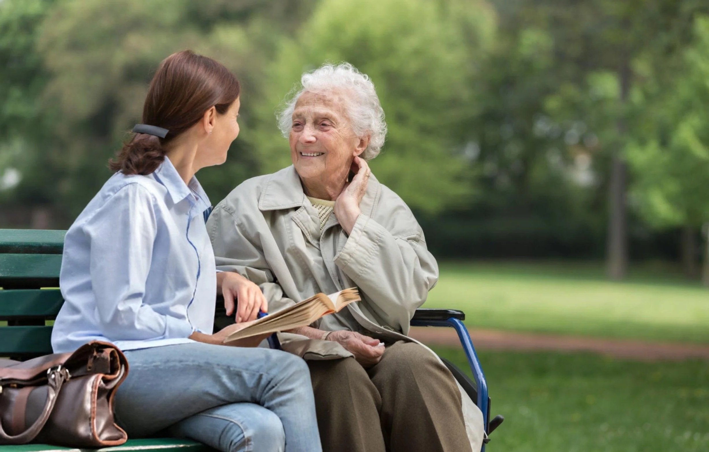 An elderly woman and a middle-aged woman sit on a park bench having a pleasant conversation, surrounded by green trees and out of focus greenery in the background.