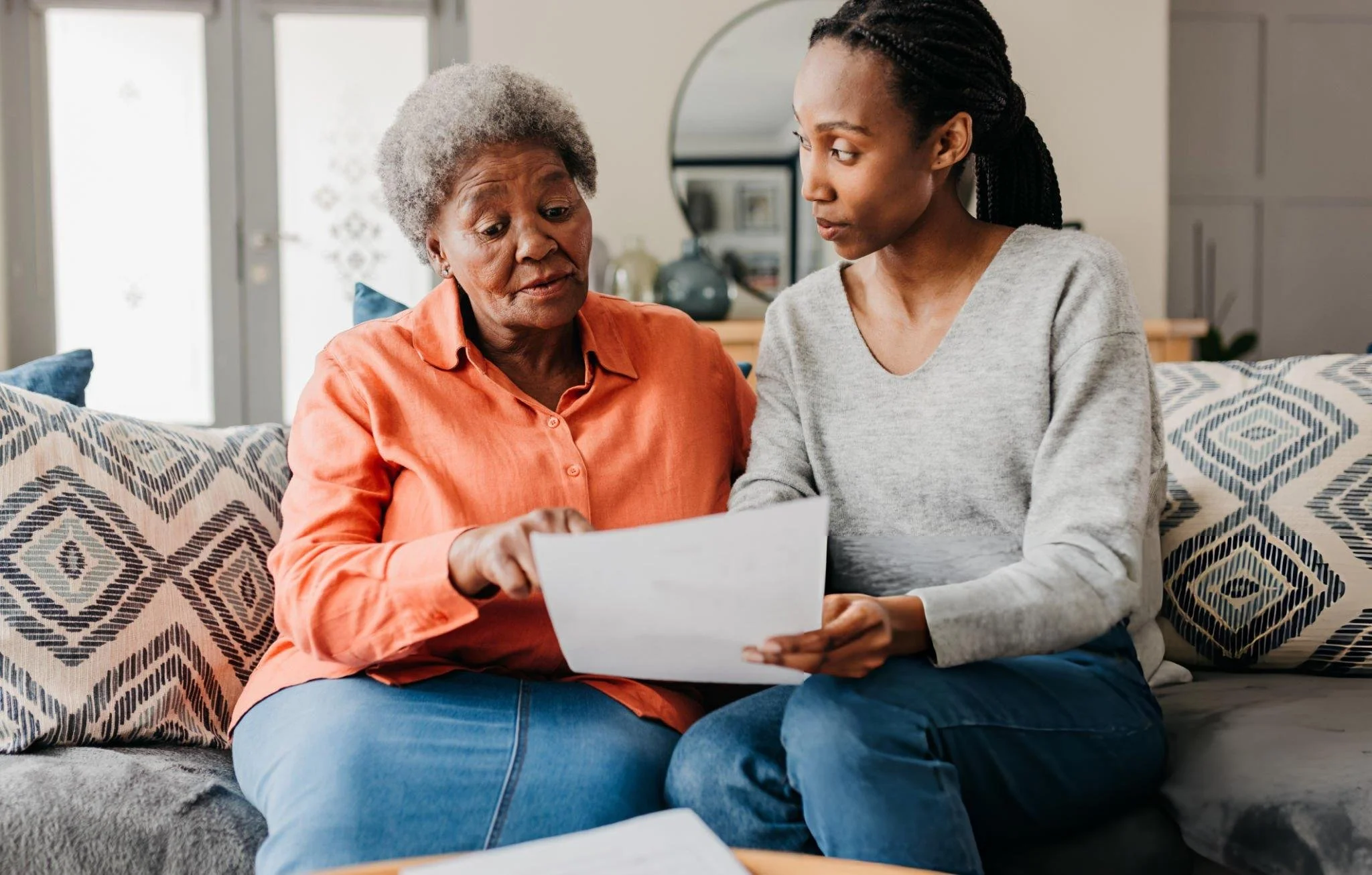 A young woman and an elderly woman sitting together on a couch, looking at a piece of paper or card.