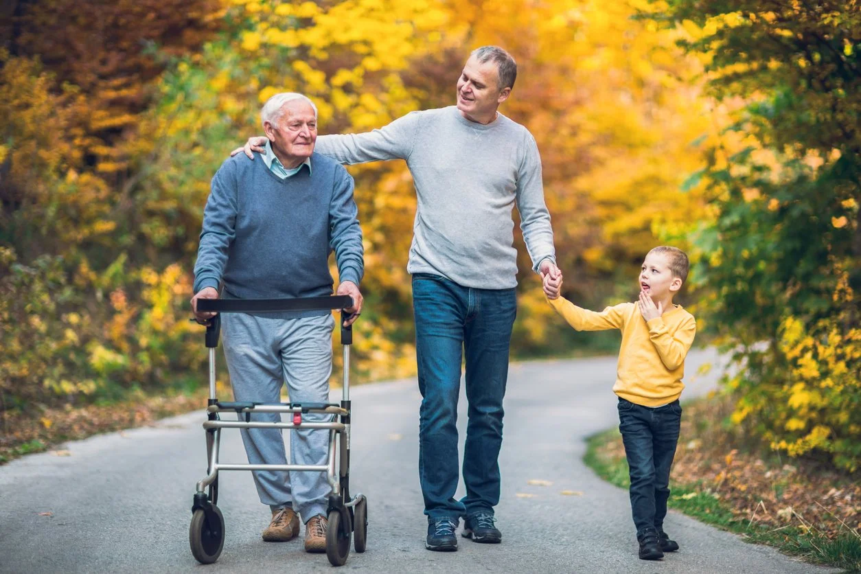 A multigenerational family walks outdoors on a path surrounded by fall foliage. An elderly man using a walker is accompanied by a middle-aged man holding his arm and a young boy holding the middle-aged man's hand.
