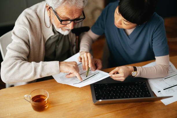 An elderly man and a young woman are sitting at a wooden table, reviewing documents together; a laptop, a cup of tea, and additional papers are also on the table.