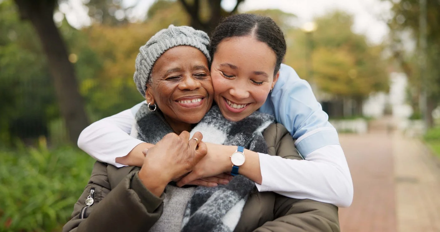Two women hugging outdoors, smiling, with trees and a pathway in the background.