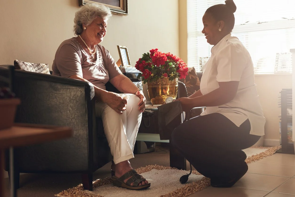 An elderly woman with gray hair smiling while sitting on a couch, talking to a nurse or caregiver who is kneeling and holding a small black bag. The room has a window with blinds, a vase with red flowers on a table, and a framed picture on the wall.