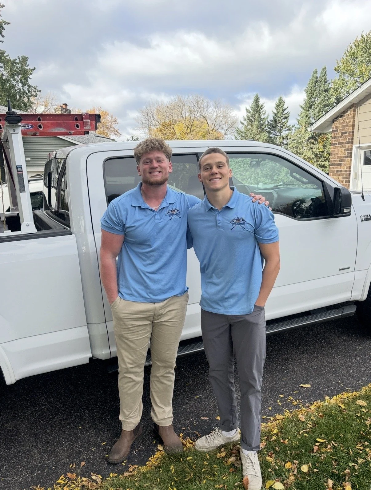 Two young men wearing blue polo shirts with logos, standing outside in front of a white truck, smiling, with trees and a house in the background.