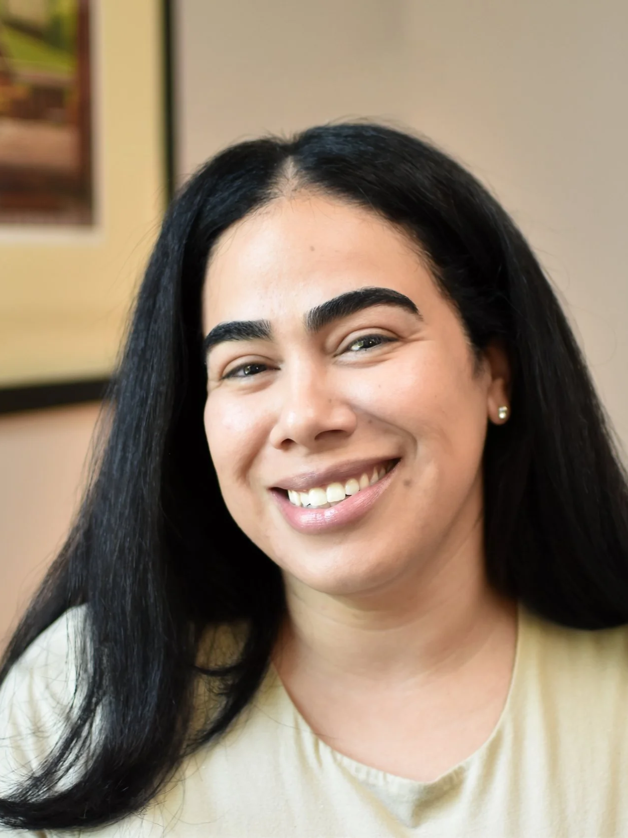 Close-up of a smiling woman with long black hair, wearing pearl earrings and a beige top.