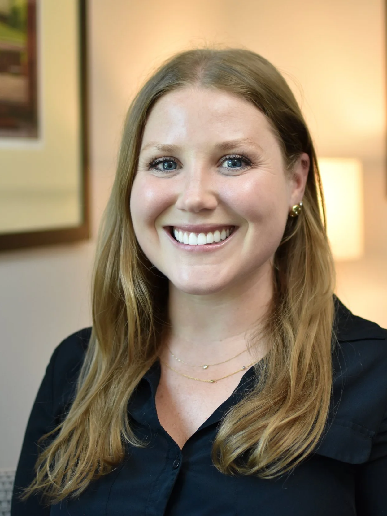 A woman with long, wavy red hair, blue eyes, wearing gold earrings, a gold necklace, and a black shirt, smiling in front of a warm-toned background with framed photos and a lamp.