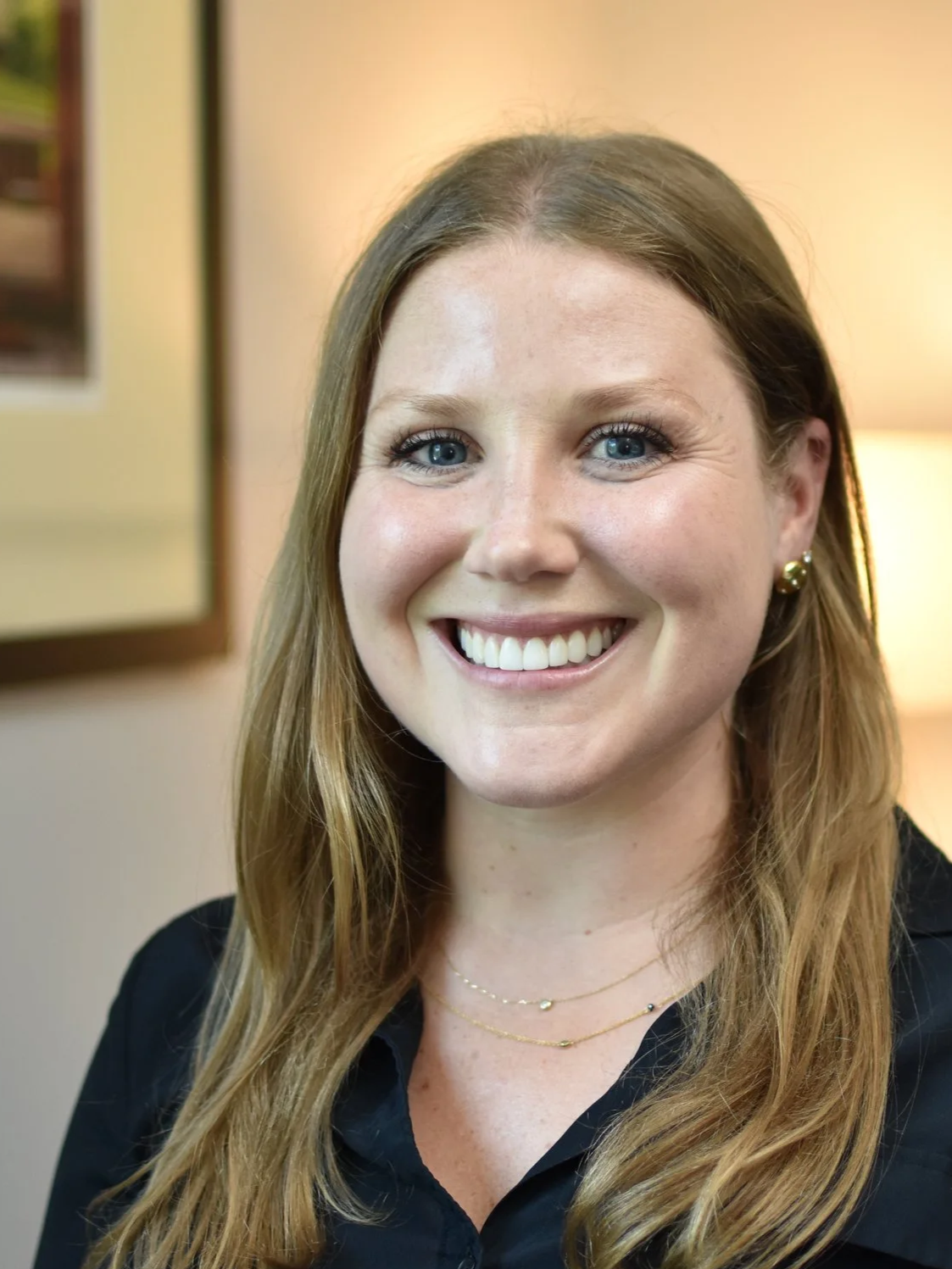 A woman with long, wavy red hair, blue eyes, wearing gold earrings, a gold necklace, and a black shirt, smiling in front of a warm-toned background with framed photos and a lamp.