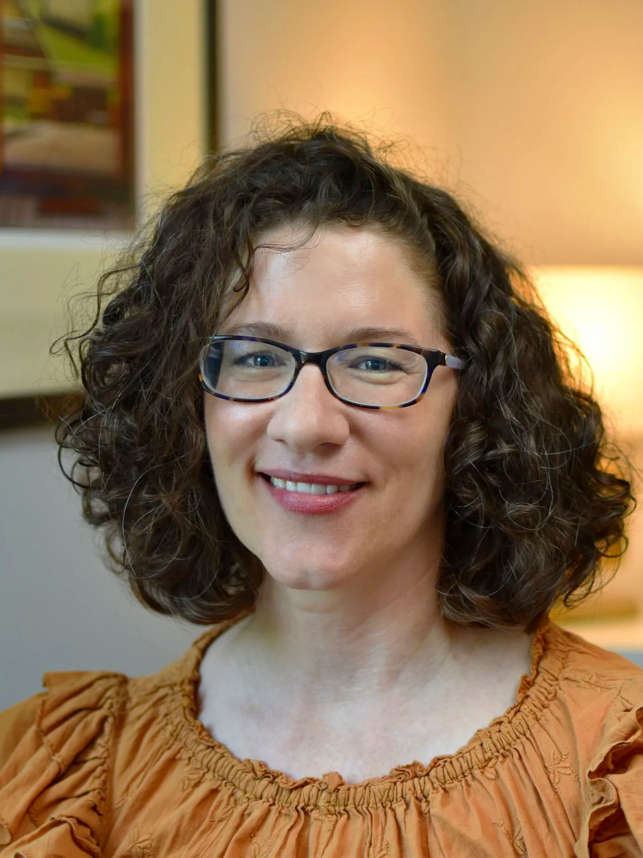 A woman with curly brown hair and glasses smiling at the camera, wearing an orange blouse, in a warmly lit room.