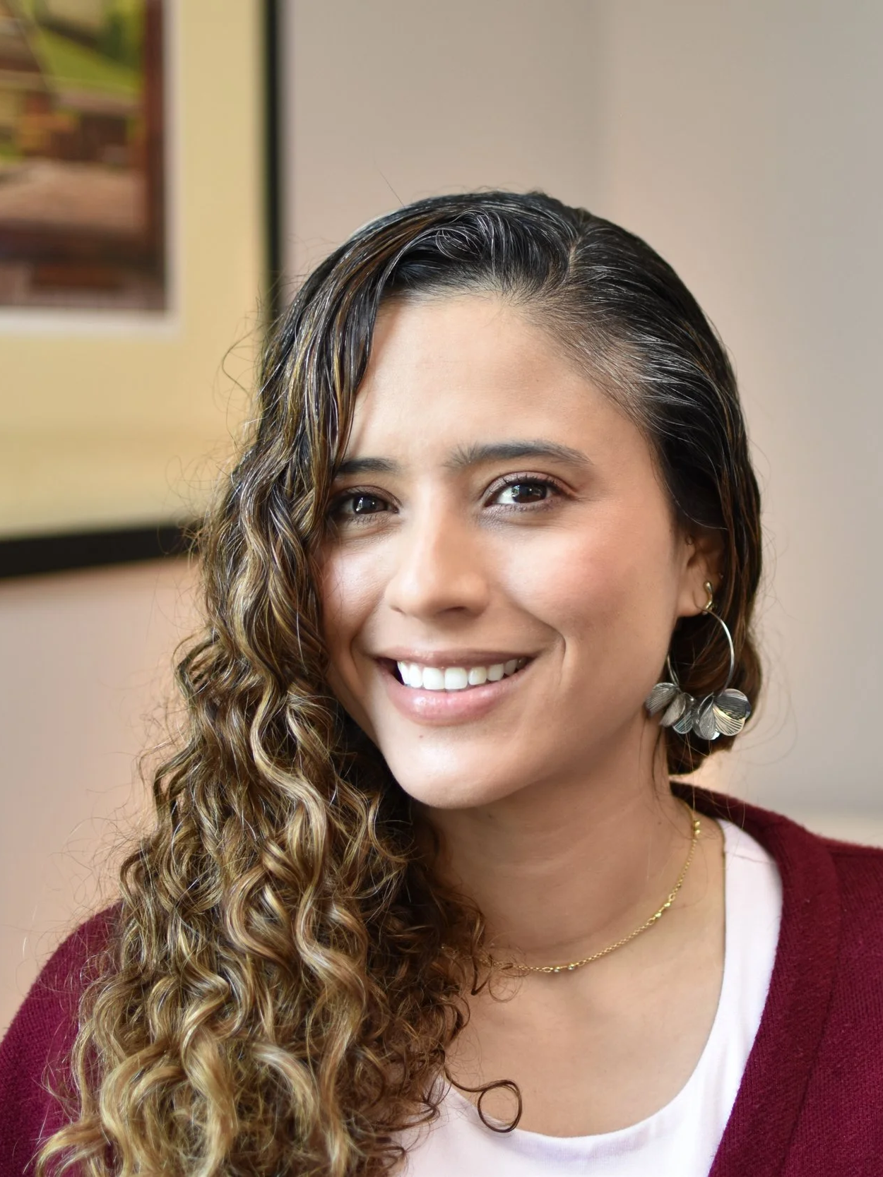 A woman with curly brown hair smiling, wearing a black blazer and a cream blouse, against a light blue background.