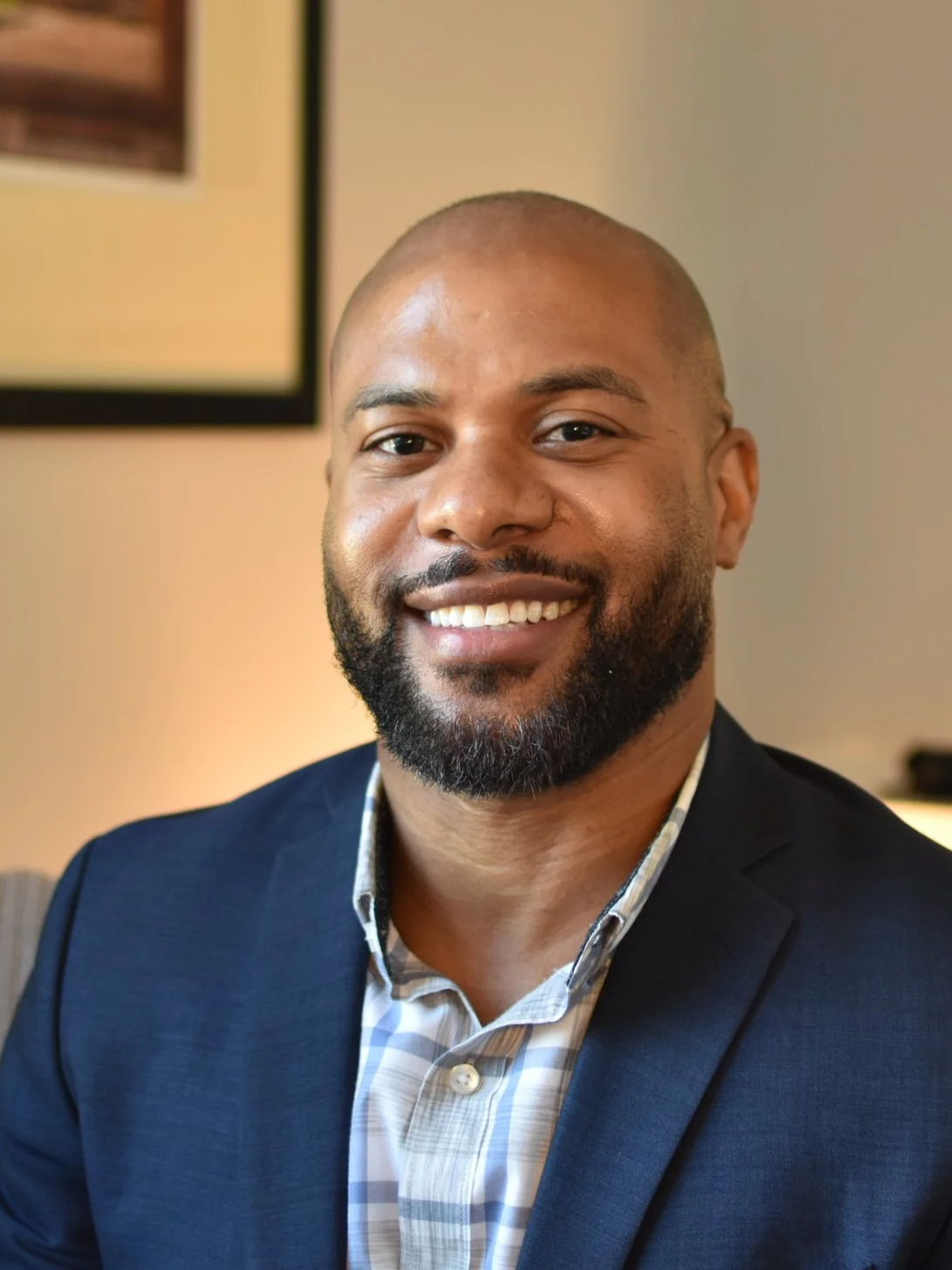 A smiling man with a beard in a navy blue suit and a plaid shirt standing indoors.