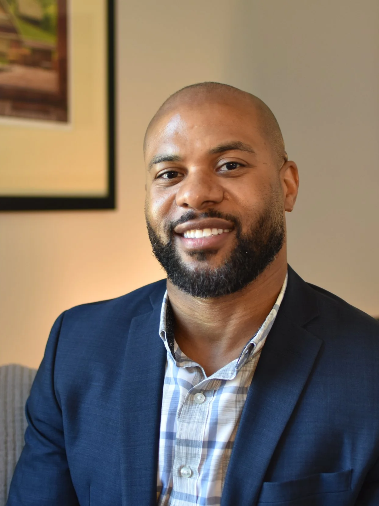 A smiling man with a beard, wearing a navy blazer and a plaid shirt, sitting indoors with a framed picture on the wall behind him.