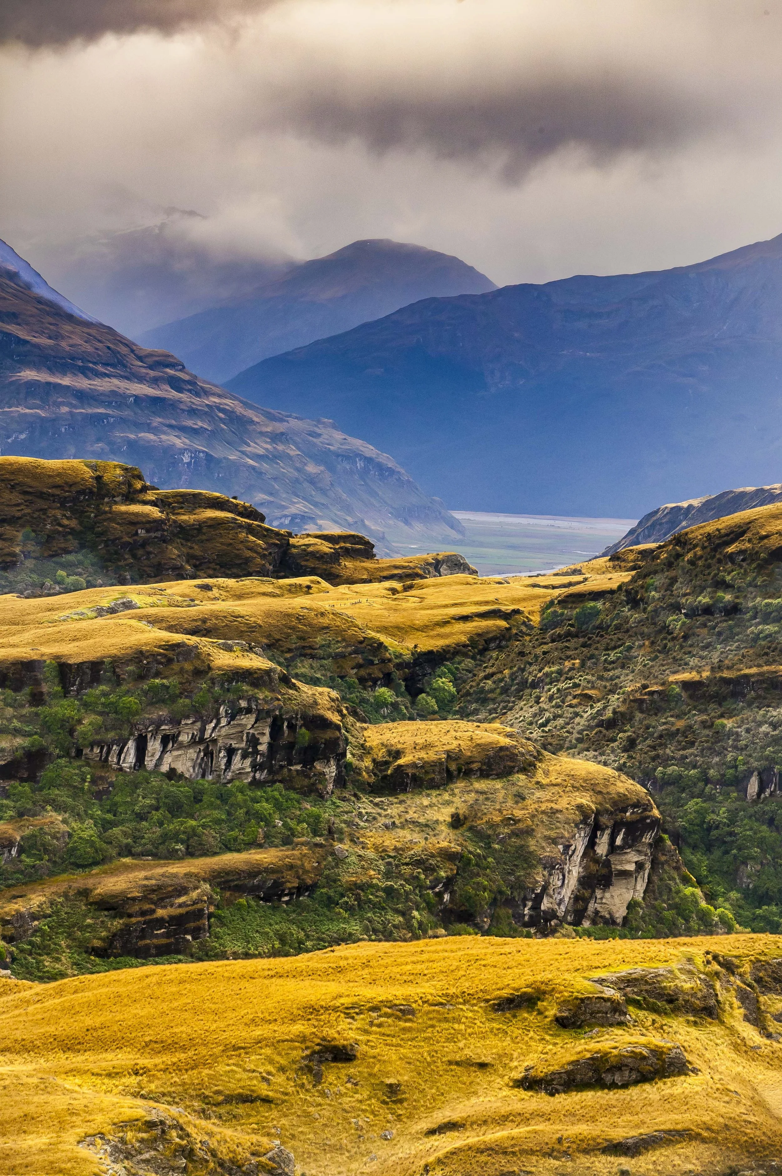 Scenic view of rolling hills with vibrant green and yellow grass, steep cliffs covered in greenery, and distant mountains under cloudy sky.