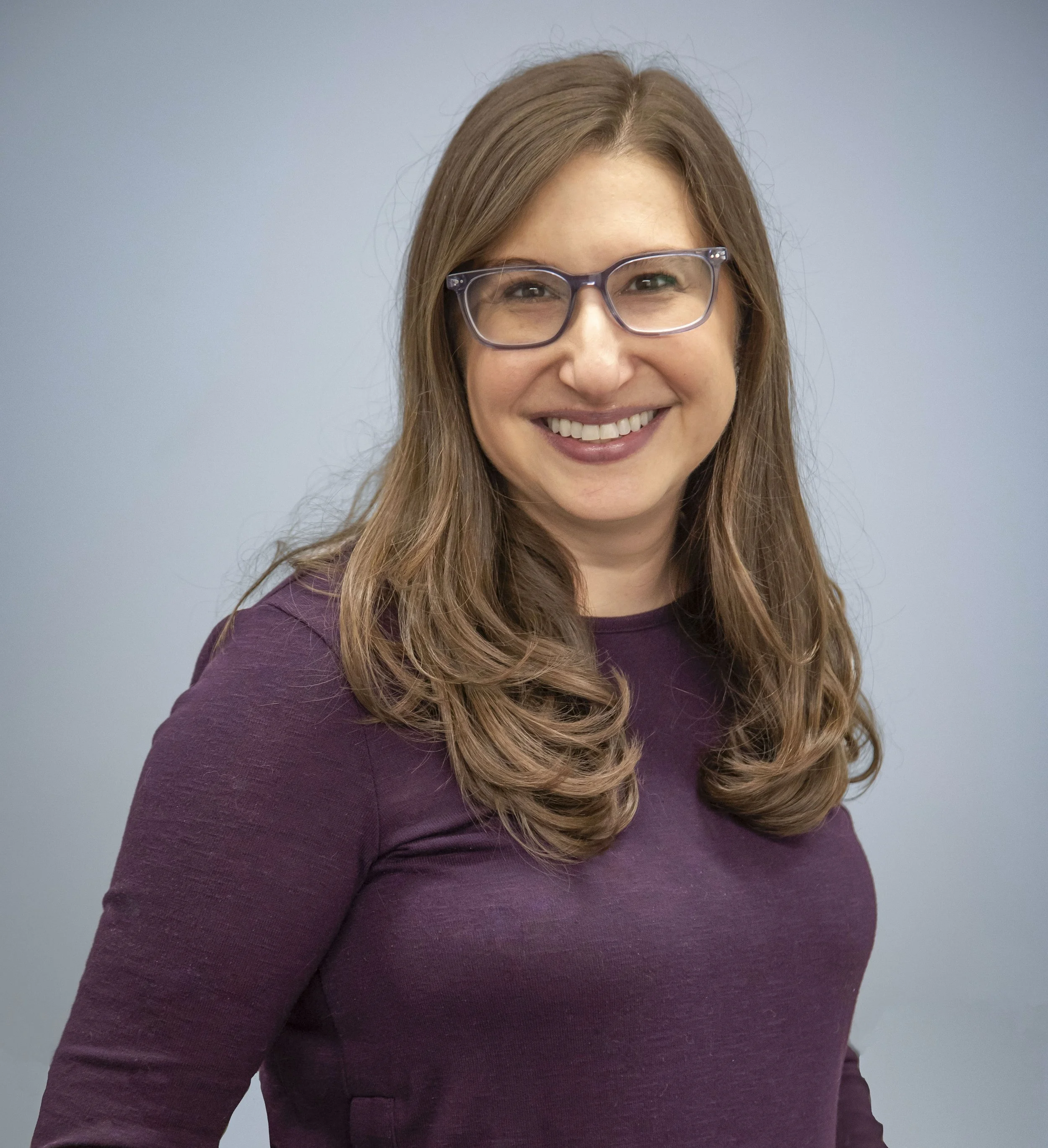 A woman smiling, wearing glasses and a purple top, standing against a plain light blue background.