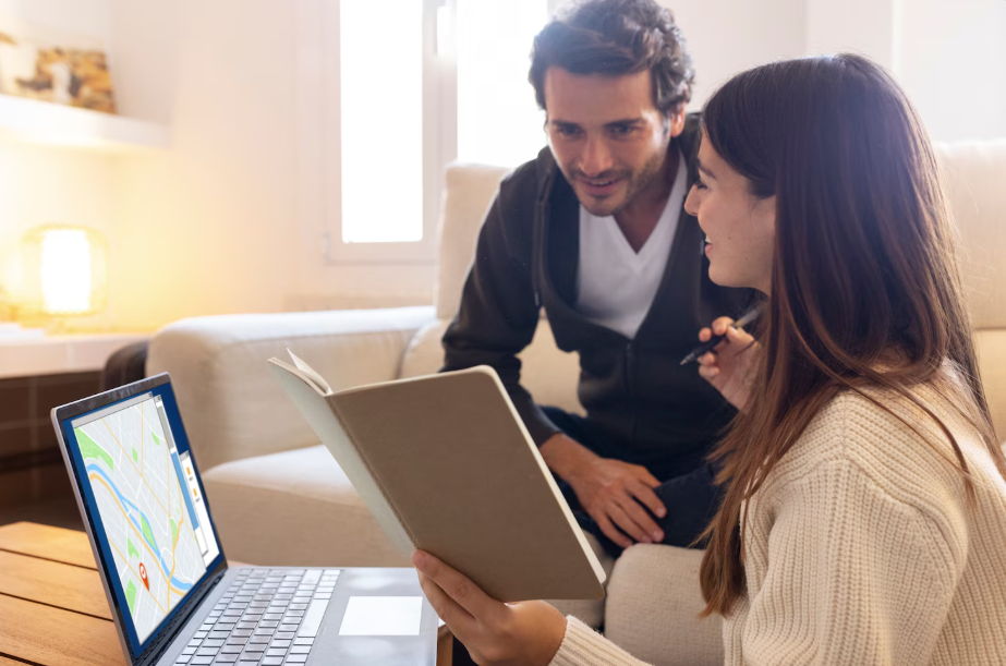 A man and woman sitting on a couch, looking at a laptop screen displaying a map, with the woman holding a notebook.