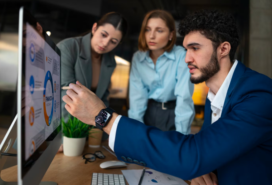 Three business professionals discussing financial data on a large computer screen in an office.