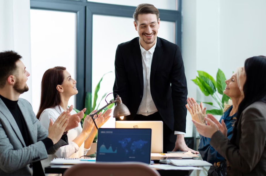 A man standing and smiling in a meeting room with four seated colleagues clapping and smiling.