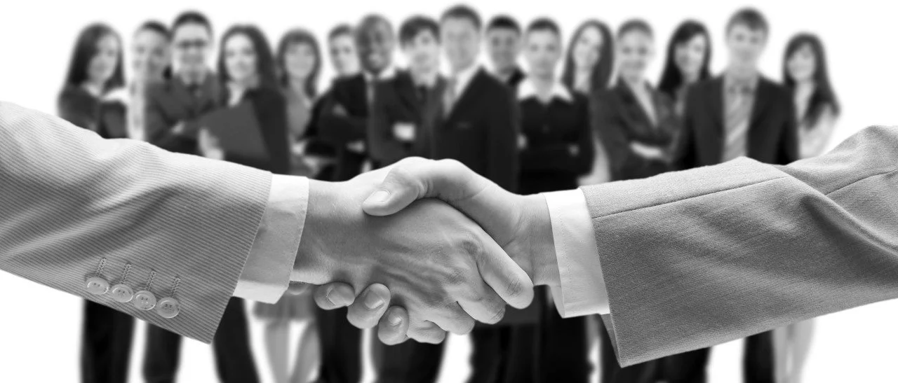 Two people shaking hands in a business setting, with a diverse group of professionals standing in the background.