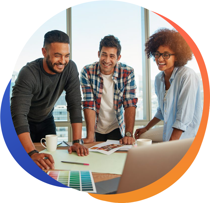 Three people are gathered around a table in a bright office, smiling and engaging in a discussion with colorful materials and documents in front of them.