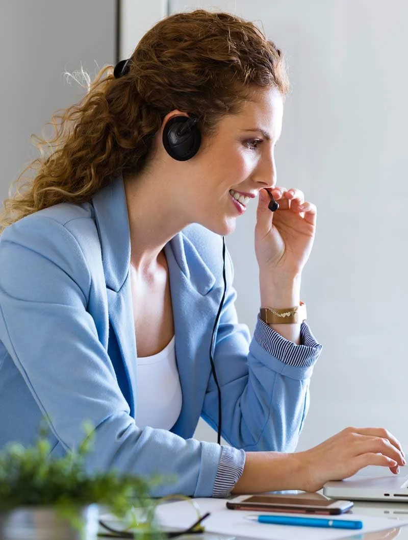 A woman with curly hair wearing a light blue blazer and white shirt is working at a desk with a computer, wearing a headset with a microphone. She is smiling, holding the microphone near her mouth with one hand and typing with the other.