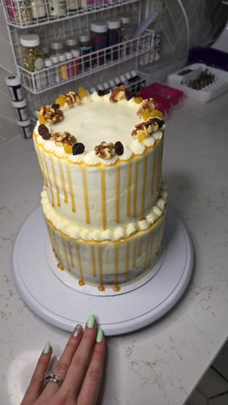 A two-tiered cake with white frosting and caramel drizzle, topped with nuts and dried fruits, displayed on a white cake stand.