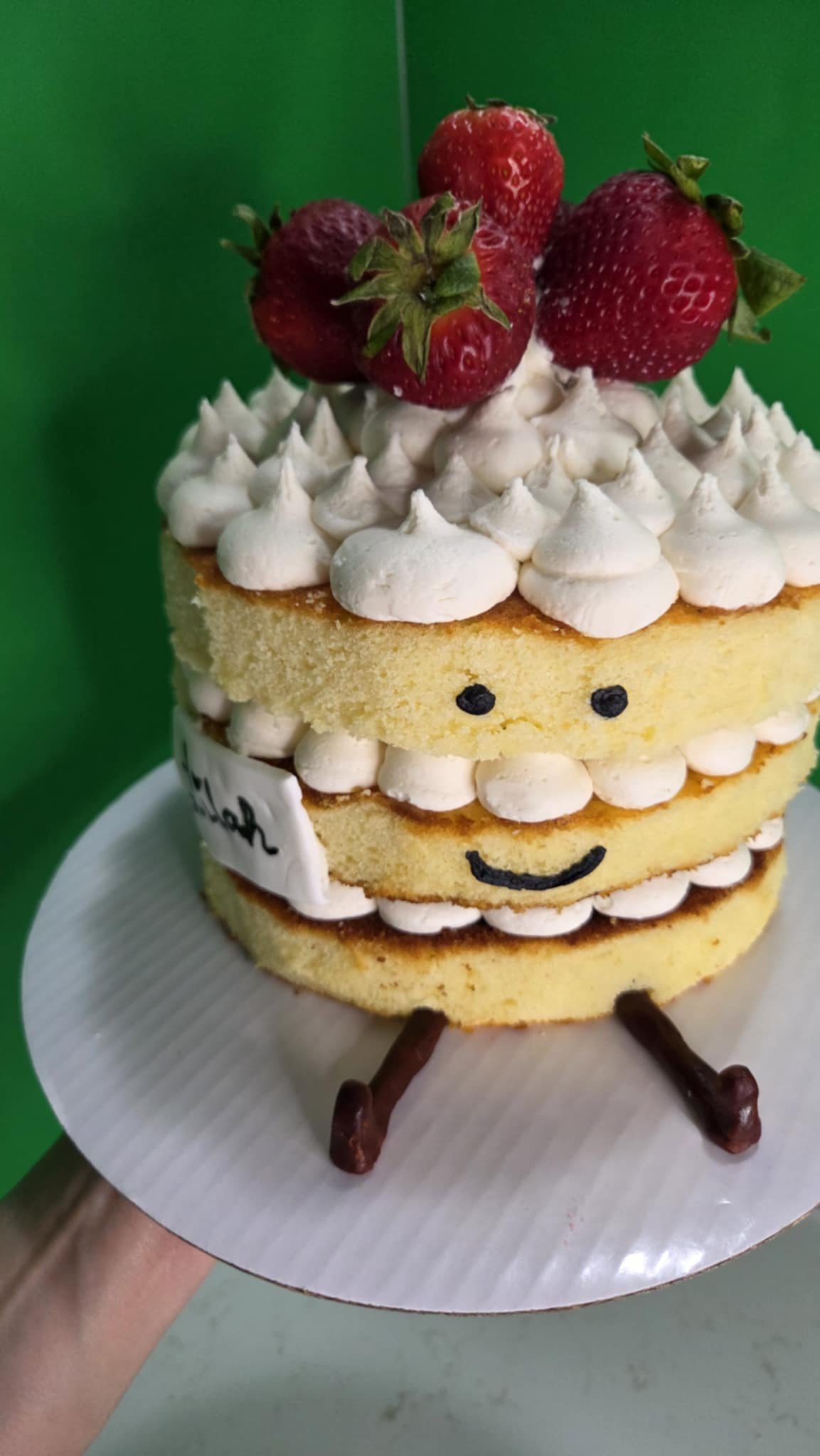 Layered vanilla cake with whipped cream frosting and fresh strawberries on top, decorated with a smiley face and chocolate sticks for legs, on a white platter.