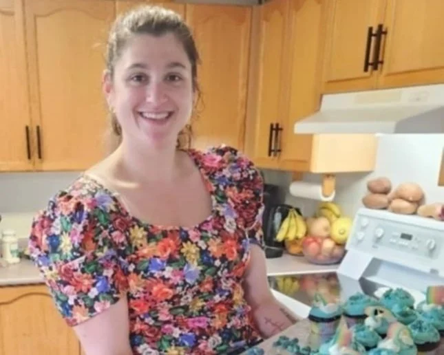 Person in floral dress holding a tray of blue cupcakes in a kitchen.