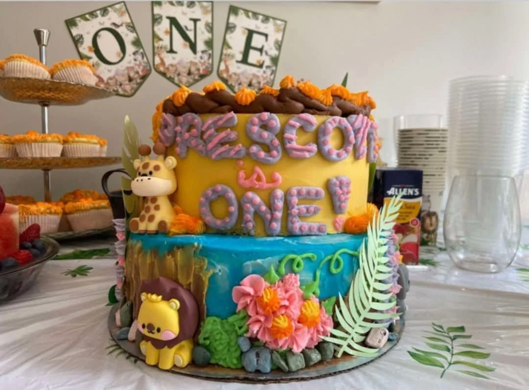 Colorful jungle-themed first birthday cake with giraffe and lion decorations, surrounded by cupcakes and a fruit platter.