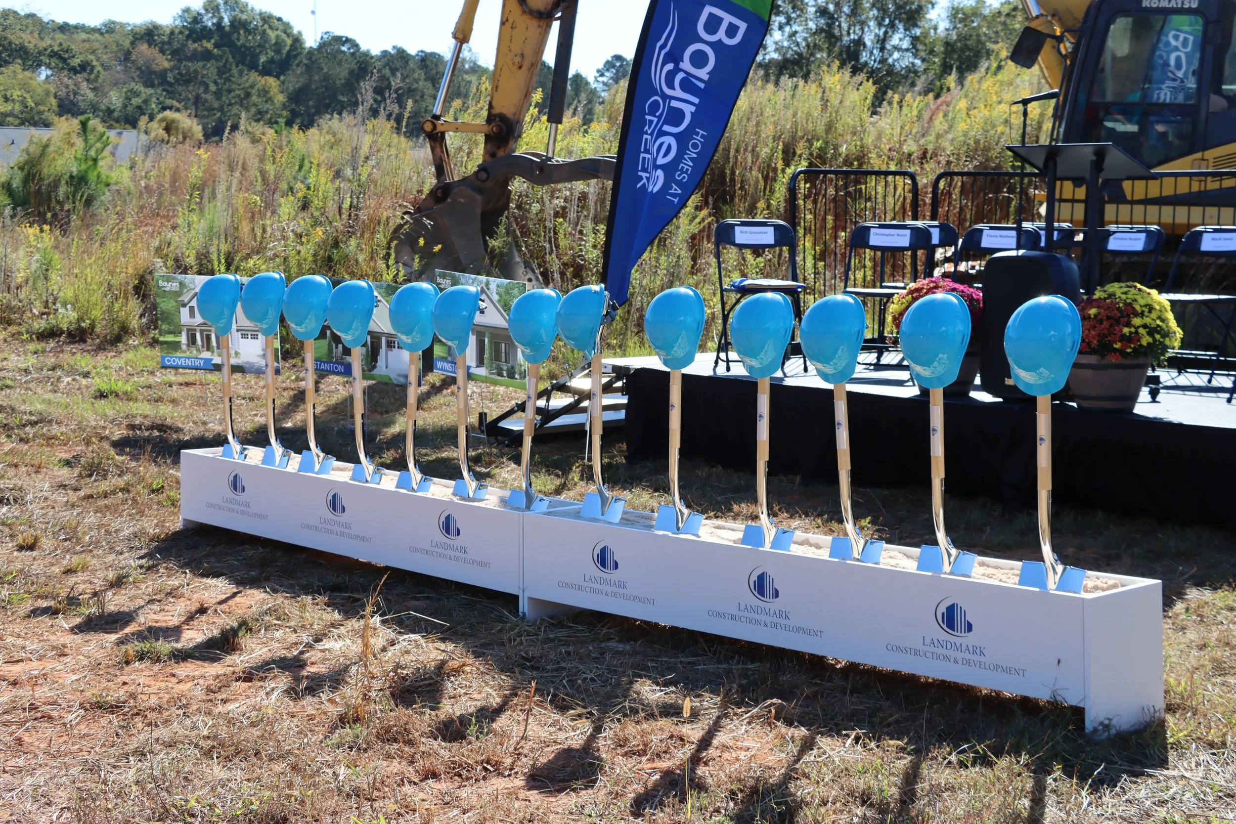 A display of small blue shovels with the Landmark Construction & Development logo, a row of blue chairs with name tags, a blue flag with white text, and a yellow excavator in the background on a grassy field at the ground breaking for Baynes Creek.