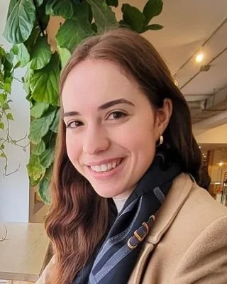 A young woman with long brown hair smiling in a well-lit indoor space, with green plants and ceiling lights in the background.