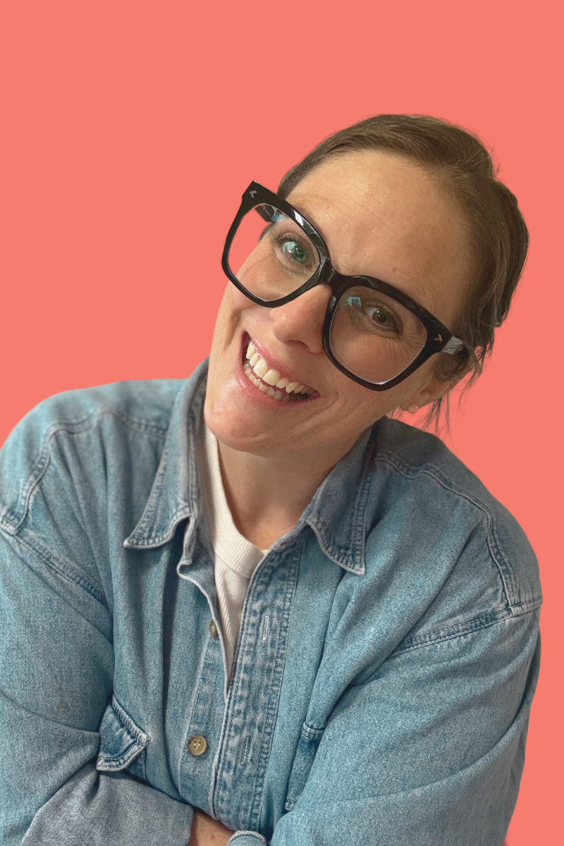 A woman with short brown hair wearing large black glasses and a denim jacket, smiling in front of a coral pink background.