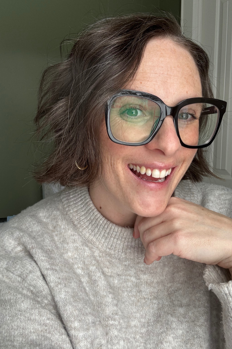A woman with short, wavy brown hair, wearing large black glasses, smiling with teeth showing, wearing a cream-colored sweater, with her hand resting under her chin.