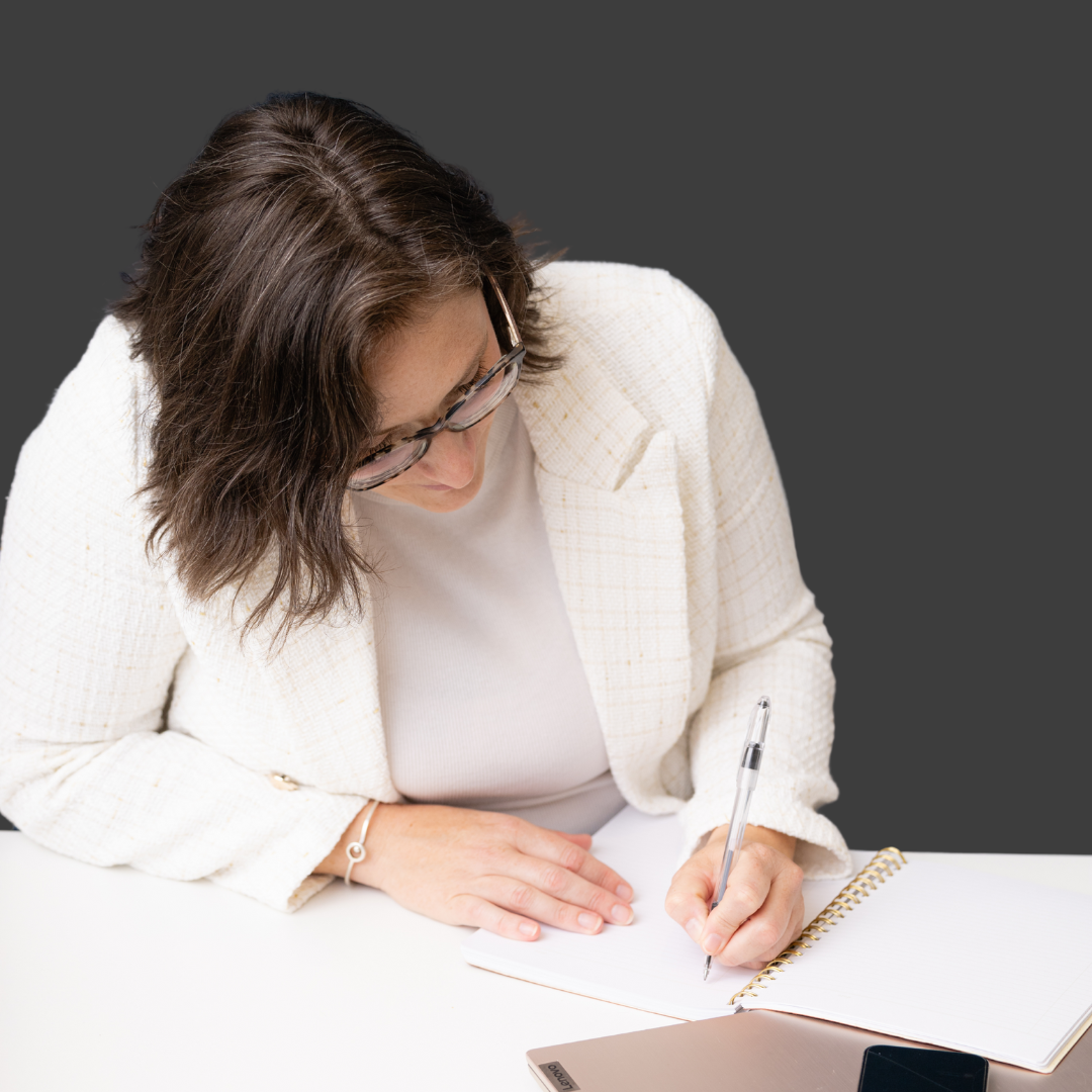 A woman with brown hair and glasses writing in a spiral notebook at a white table with a silver laptop and black phone nearby.