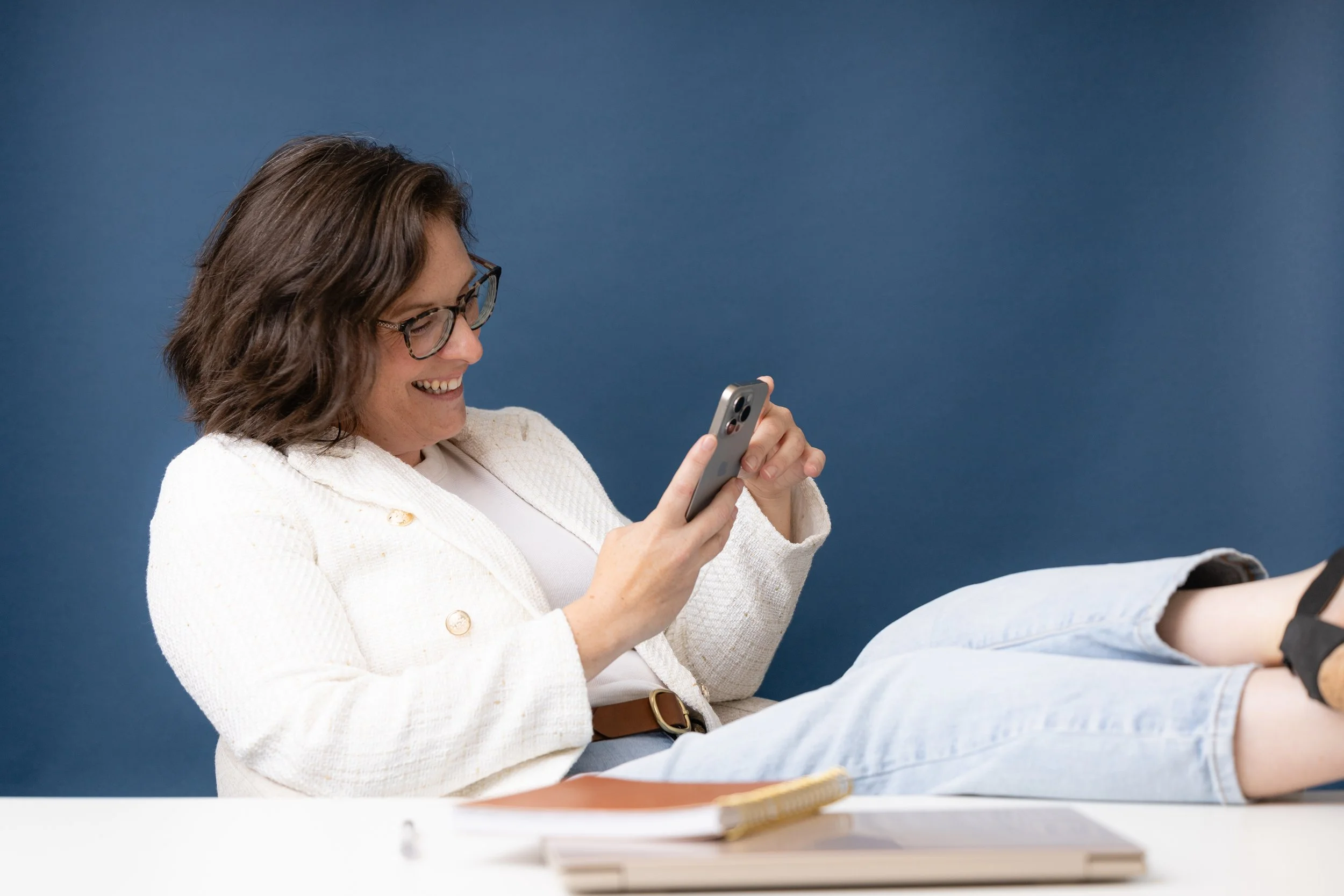 Woman with glasses smiling and using a smartphone, sitting with her legs stretched out on a desk against a blue wall.