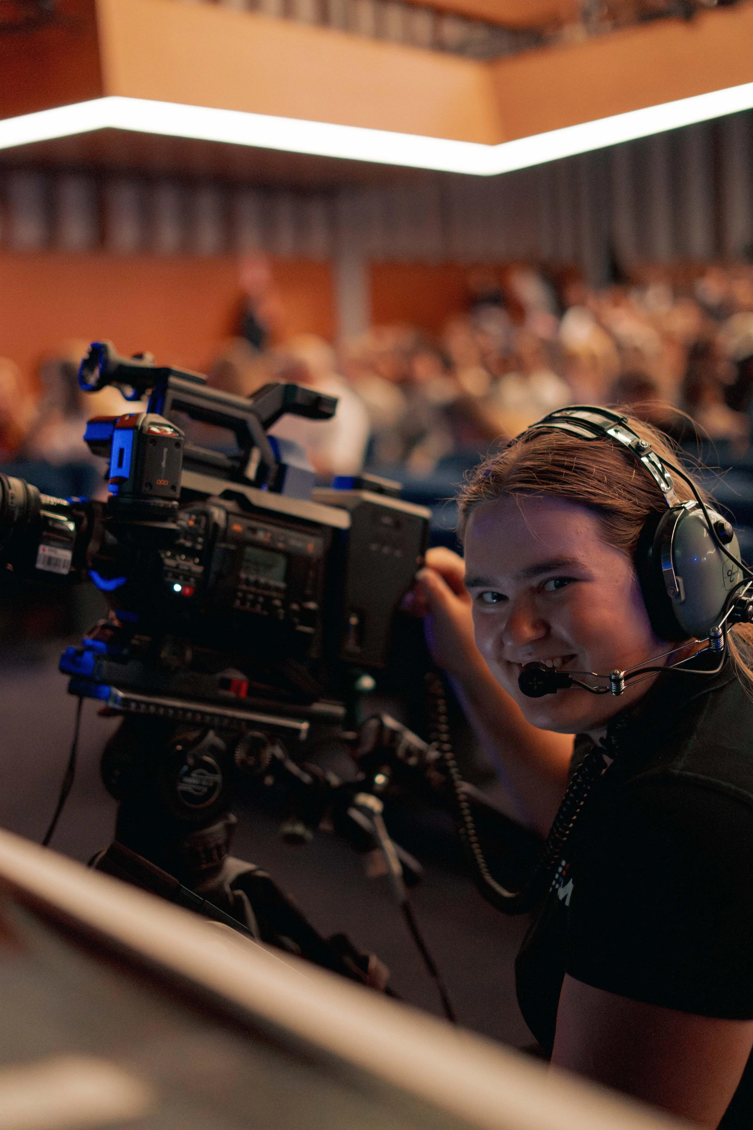 A person operating a professional camera on a tripod in a crowded indoor setting, wearing headphones, with a head full of reddish-brown hair, smiling and looking towards the camera.