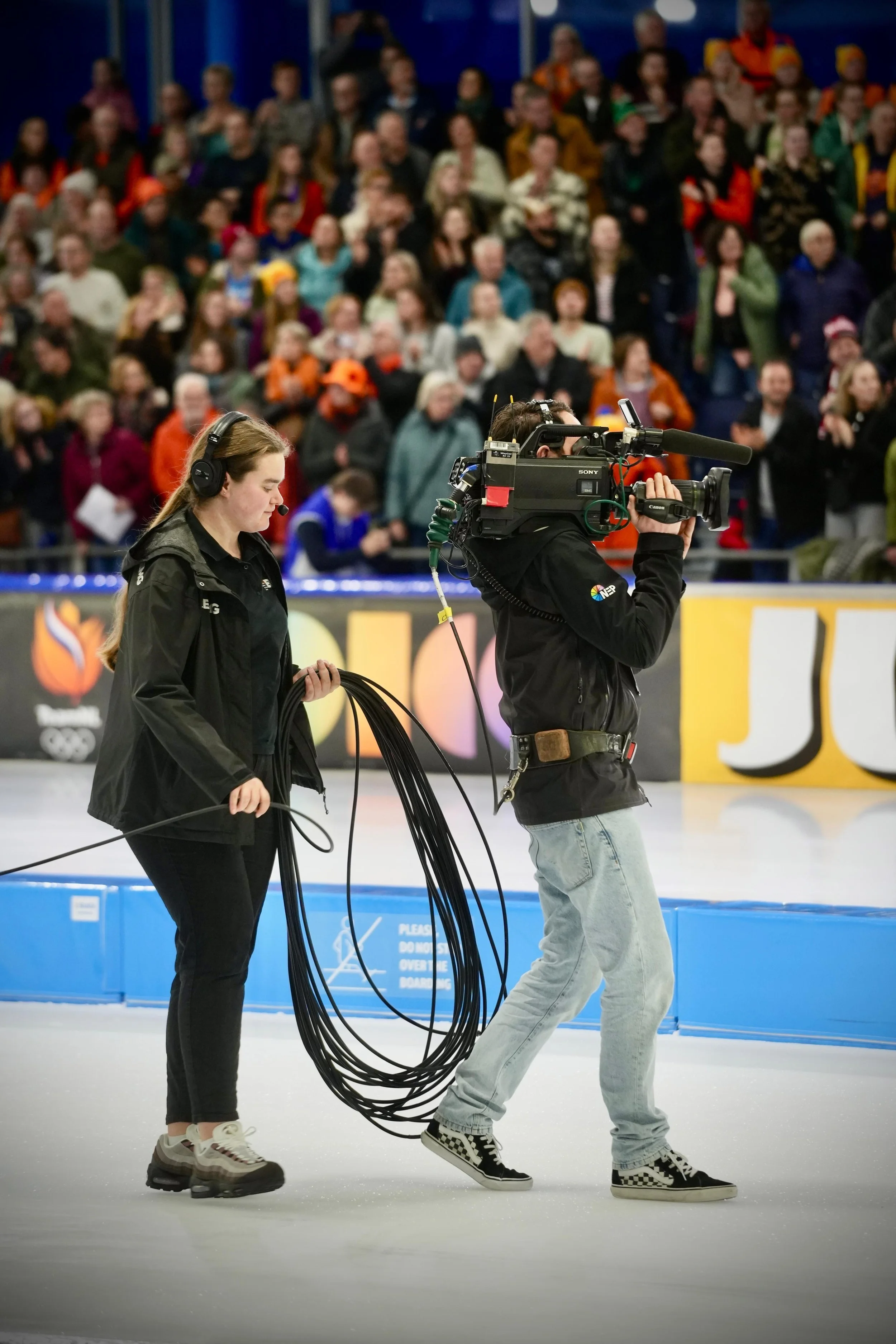 Two camera operators filming on an ice rink during the Olympics, with a large crowd watching in the background.
