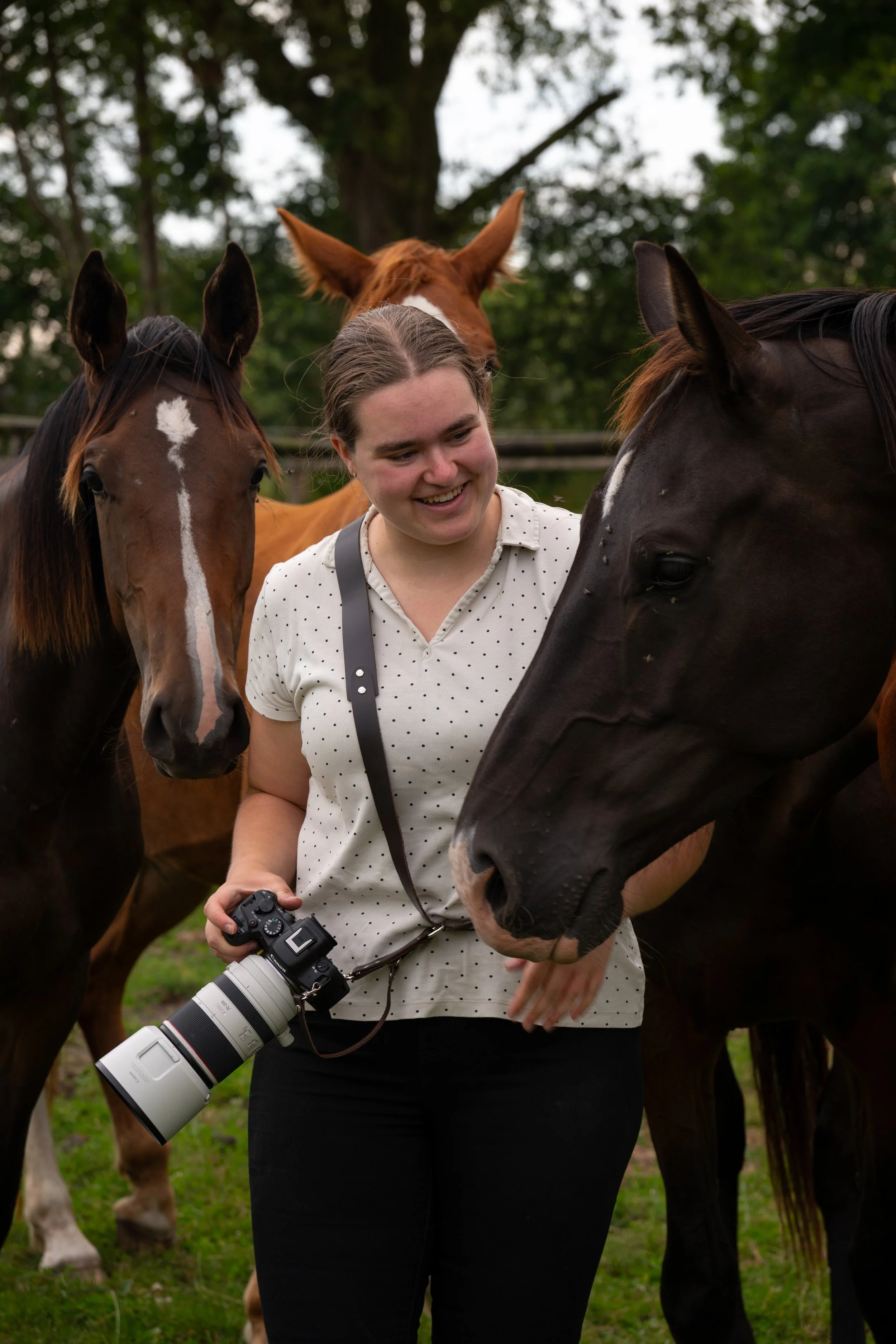 A woman in a white polka dot shirt and black skirt smiles as she interacts with a black horse, holding a camera with a large lens; two other horses, one brown and one chestnut, stand nearby in a grassy outdoor setting with trees in the background.