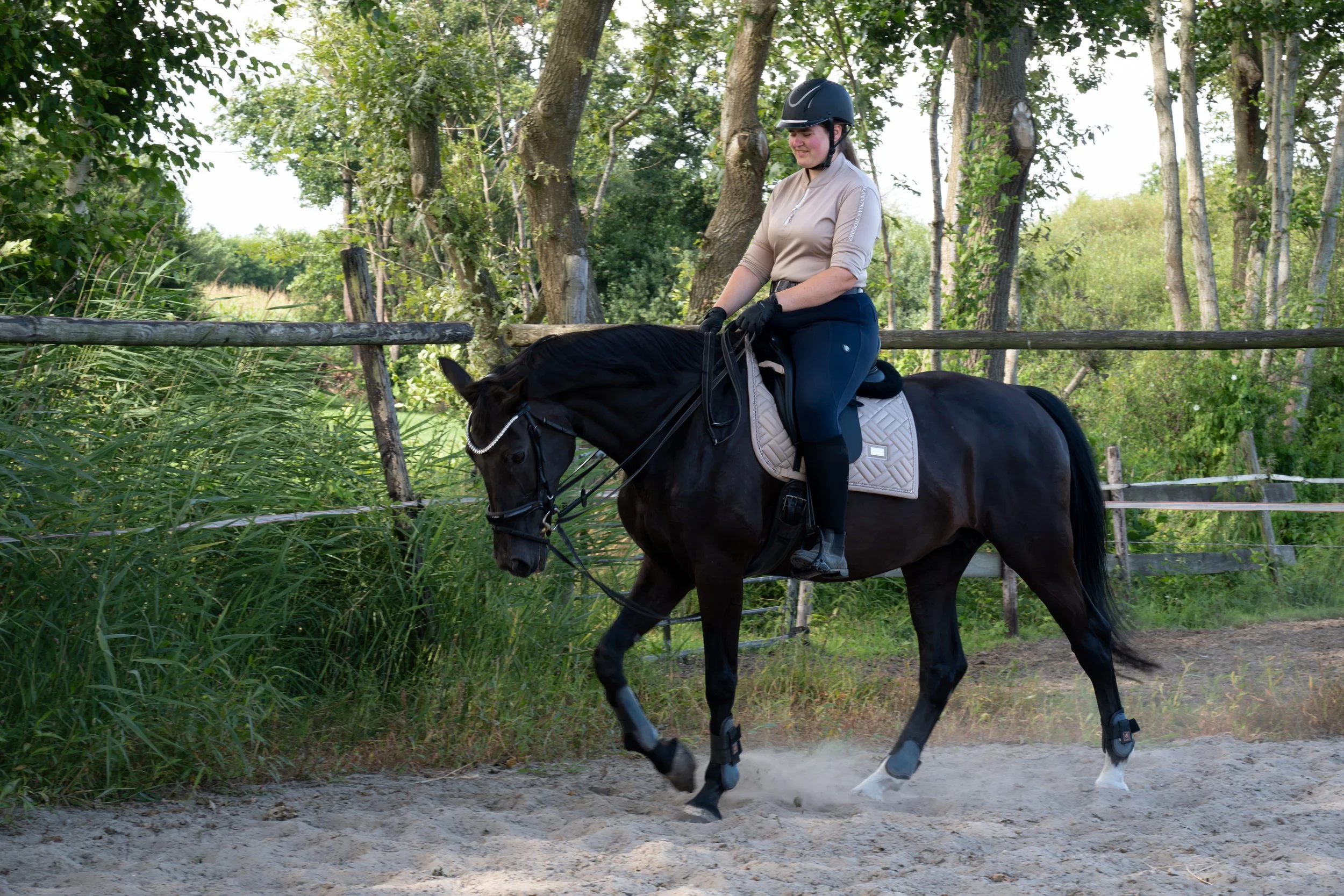 A woman riding a black horse along a forest trail, wearing a beige riding jacket, riding helmet, and black gloves.