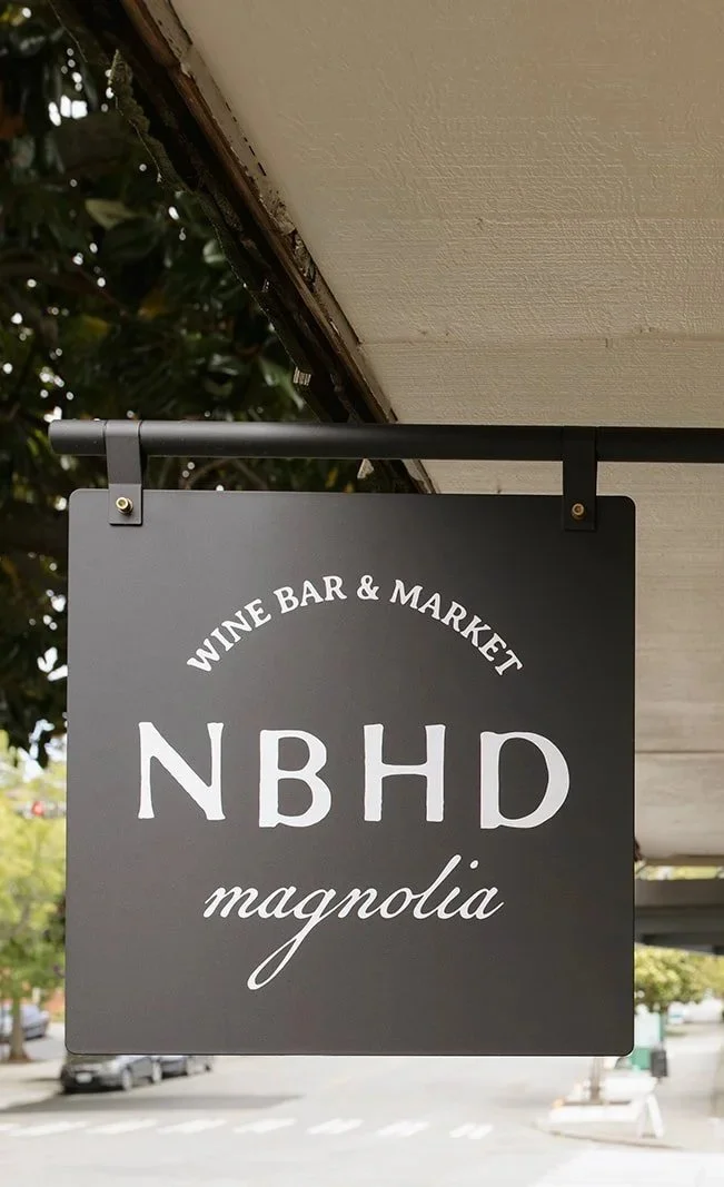 A black square sign hanging outside a building that reads "WINE BAR & MARKET NBHD magnolia" in white text.
